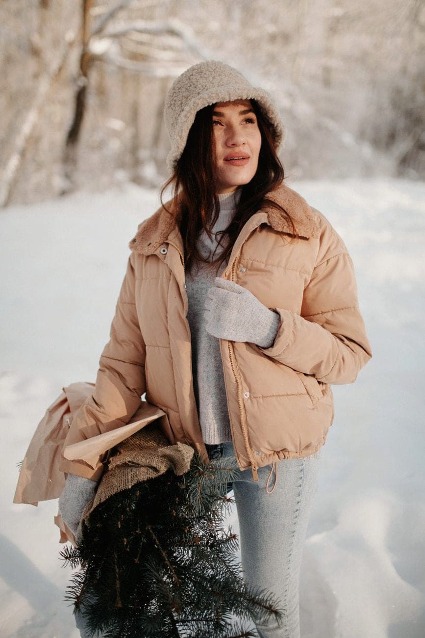 Vrouw in een camelkleurige donsjas, grijze trui en beige muts in een besneeuwd bos