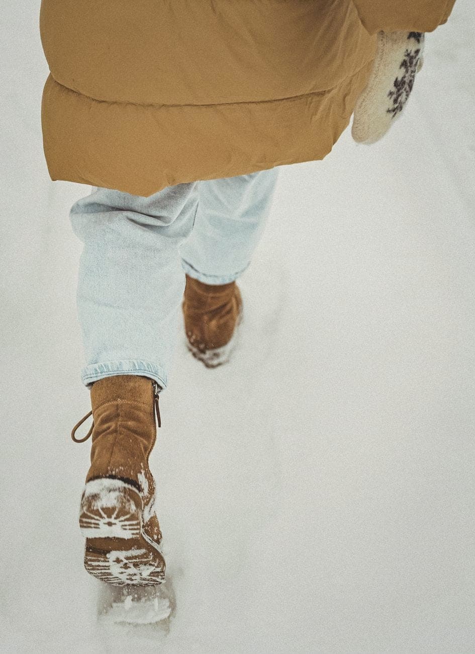 Iemand in een camelkleurige jas en veterboots die door de sneeuw stapt