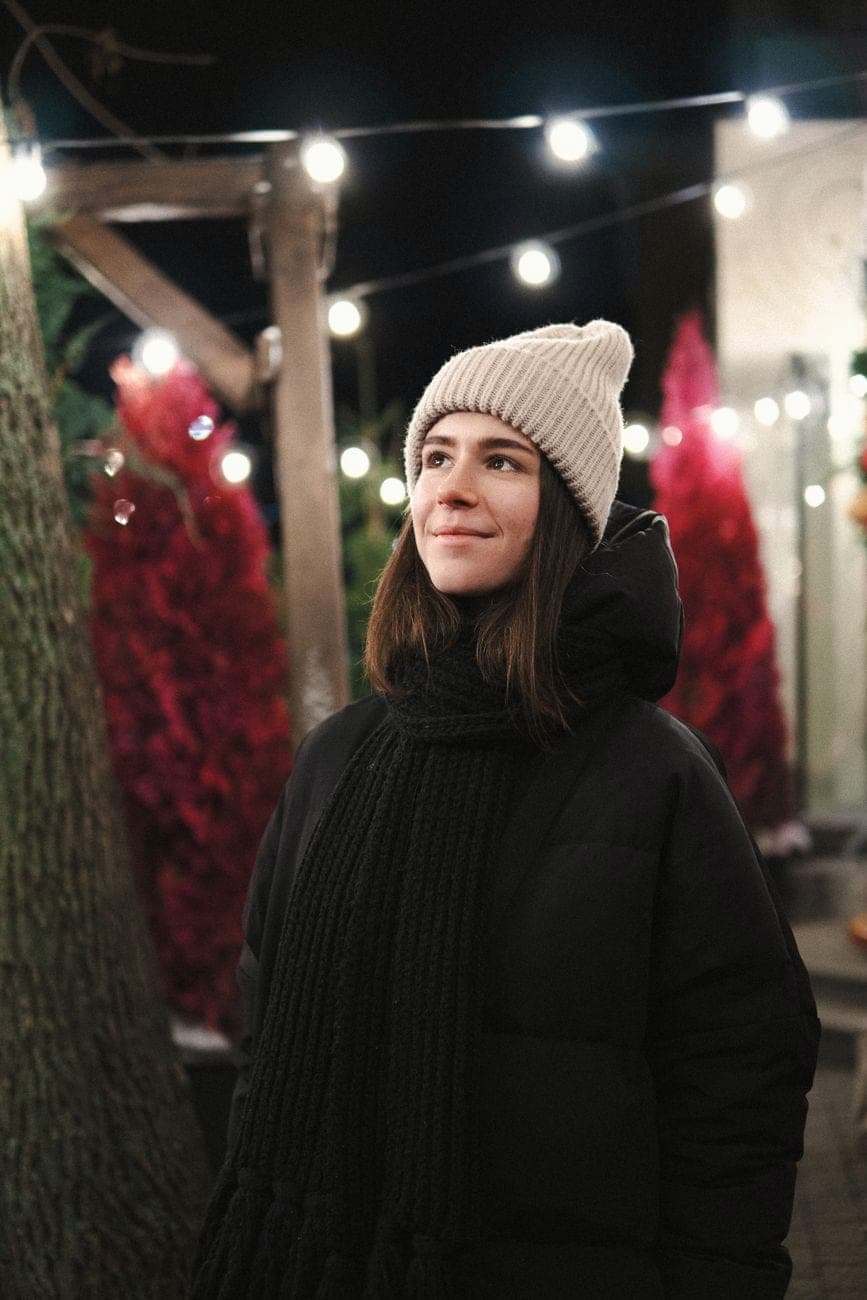 Vrouw in een zwarte donsjas en crèmekleurige muts tussen feestverlichting 's avonds