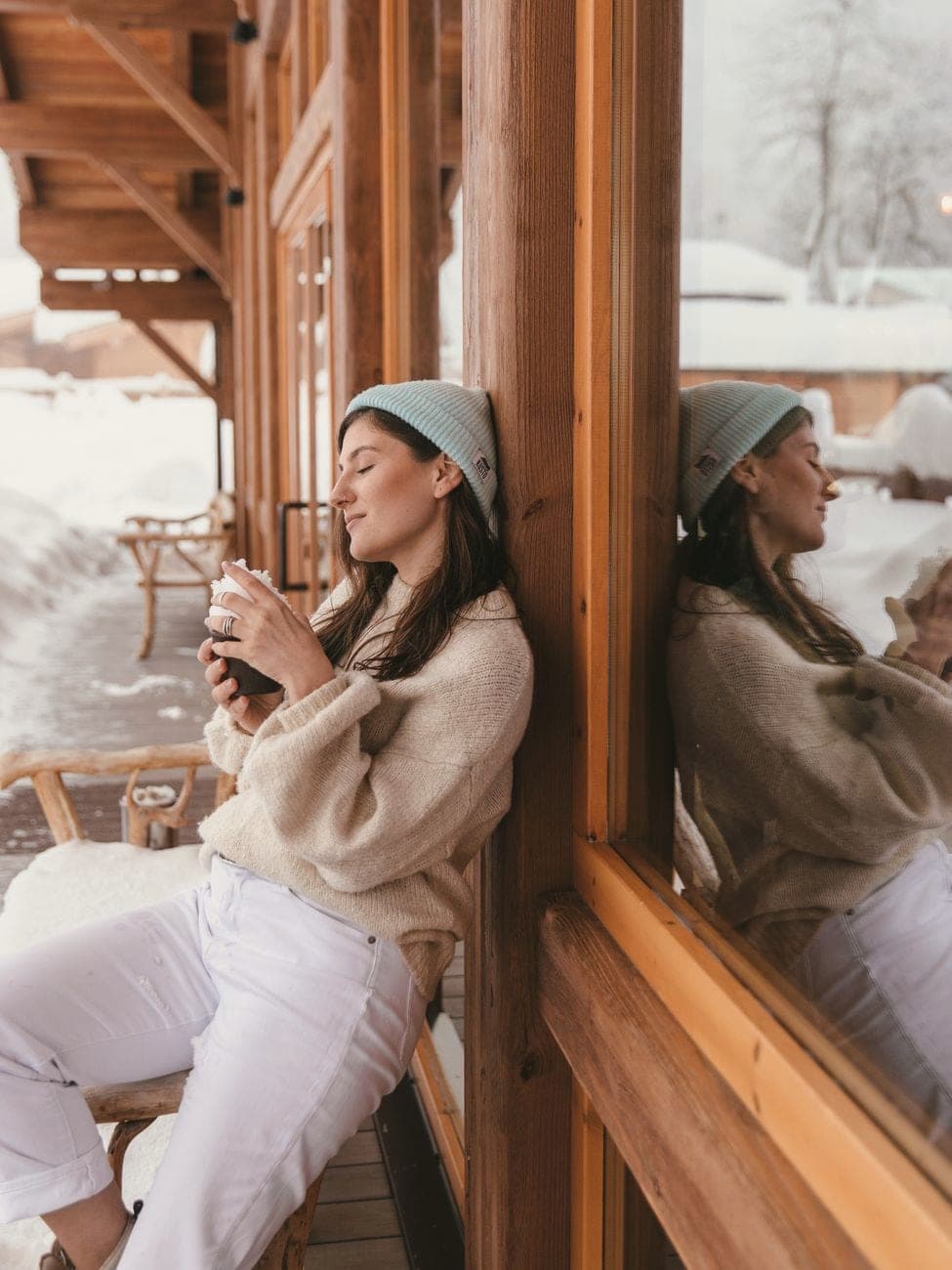 Vrouw in een crèmekleurige gebreide trui en muts tegen een houten chaletwand in de sneeuw